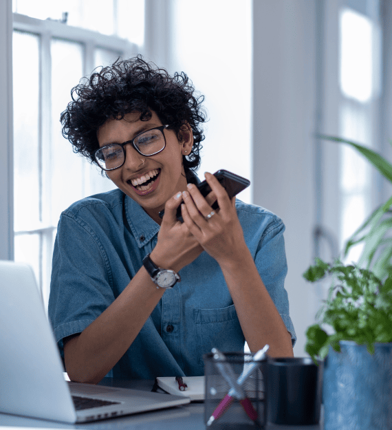 Female or non-binary presenting person looking at laptop and laughing while holding smartphone